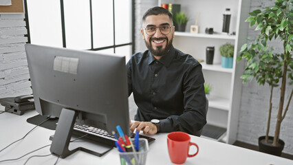 A smiling, bearded man working at his computer in a modern office setting.