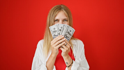 Attractive young woman with blonde hair covering mouth with us dollars against red background.