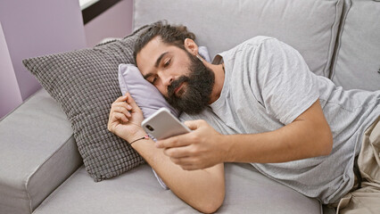 Fototapeta premium Hispanic man lounging with smartphone in cozy home interior, embodying relaxation and modern living.