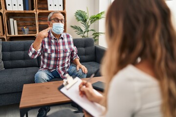 Cheerful mature man with white hair, happily pointing with his hand and smiling behind his medical mask at psychology clinic