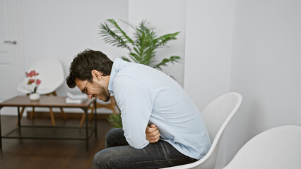 Fototapeta premium A pained young hispanic man clutches his stomach in a modernly furnished white waiting room, evoking a sense of urgency.