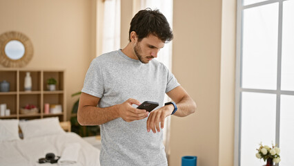 A young hispanic man checks time on his watch while holding a smartphone in a bright bedroom.