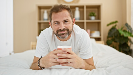 Hispanic man with grey hair relaxes with a mug in a bright, cozy bedroom, exuding a sense of home comfort and maturity.