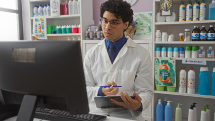 A young hispanic man working as a pharmacist in a well-stocked pharmacy, wearing a white lab coat and glasses, using a computer and taking notes in a bright, organized interior.