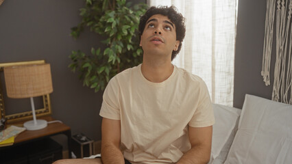 Young hispanic man sitting on a bed in a cozy bedroom, looking up thoughtfully, surrounded by home decor and indoor plants, suggesting a relaxed and introspective moment at home.