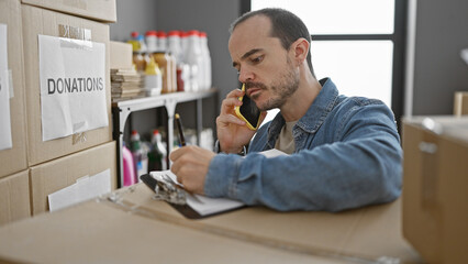 Bald bearded man working in a donation center, talking on the phone and writing notes indoors.