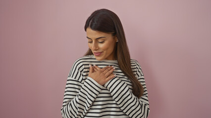 Fototapeta premium Serene hispanic woman with hands on chest, standing against a pink wall, displaying a gesture of gratitude.