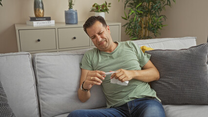 Middle-aged man indoors looking at thermometer in living room expressing concern or discomfort while sitting on couch surrounded by plants and home decor