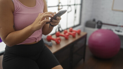 A young hispanic woman checking her phone in a gym with red dumbbells and a pink exercise ball in the background, highlighting an indoor workout setting.