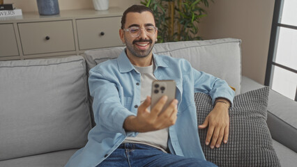 Young man with a moustache smiling at his phone while lounging on a sofa in a cozy living room with...