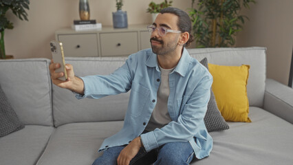 Young man with a moustache taking a selfie in the living room of an apartment, wearing glasses and a light blue shirt, sitting on a couch with yellow pillows and indoor plants in the background