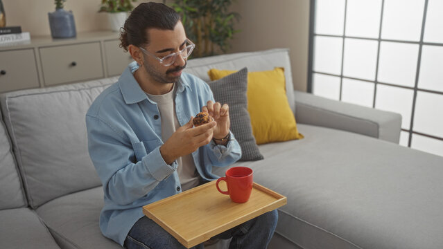 Young man with moustache eating pastry on sofa with red mug in modern living room