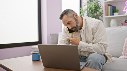 A mature hispanic man with a beard multitasking with a phone and laptop in a cozy living room.