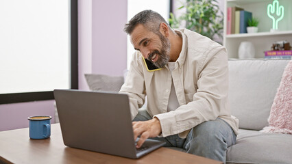 Handsome senior hispanic man with beard enjoying a conversation on his phone while seated on a sofa...