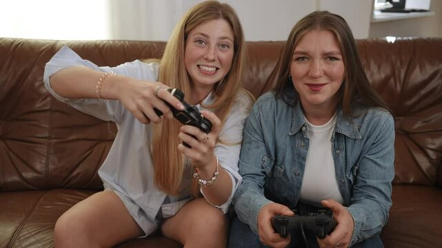 Two girlfriends look into the camera holding gamepads and quickly pressing buttons with their fingers, while sitting on a leather couch in the living room. The joy of victory and the bitterness of