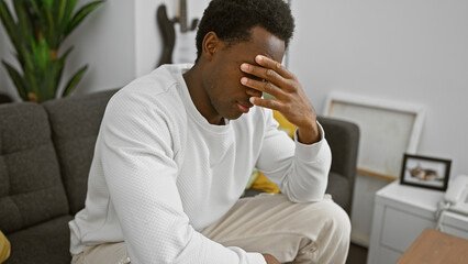 A distressed young african american man sitting indoors in a modern living room covering his face with his hand.