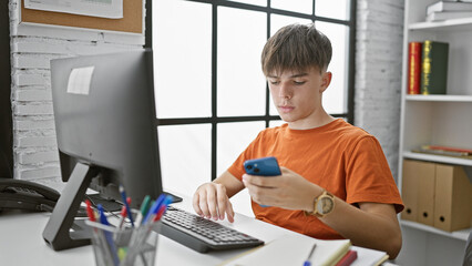 A young caucasian male teenager studies while using a smartphone and computer in a modern classroom...