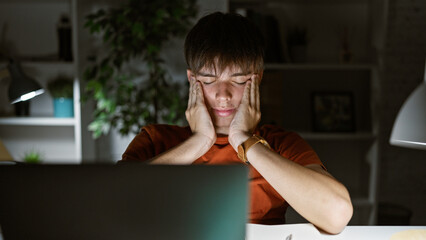 Stressed teenager man holding face in hands at computer in dark home room late night, showing...
