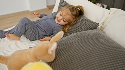 A joyful blonde little girl in pajamas lying on a bed with a stuffed toy in a cozy bedroom interior at home.