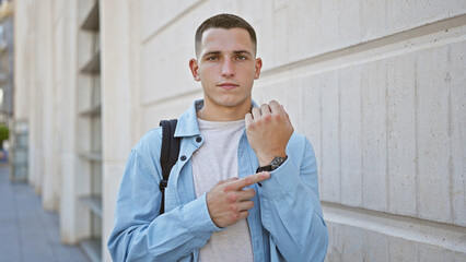 Hispanic young man in casual attire pointing at his watch on a city street, looking punctual and ready.