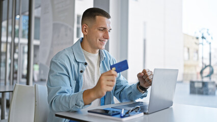 Smiling young man with credit card using laptop at university campus library.