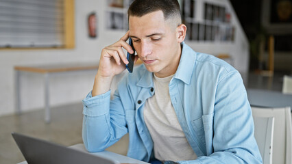 A focused hispanic man talks on a smartphone while working on a laptop in a university library.