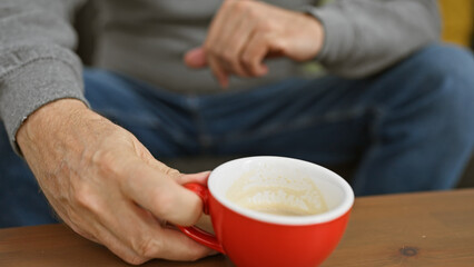 A middle-aged man relaxes at home, holding a red coffee cup at a wooden table in a comfortable interior setting.