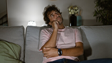 Fototapeta premium Pensive hispanic man sitting on a couch in a cozy living room, contemplating alone at night.