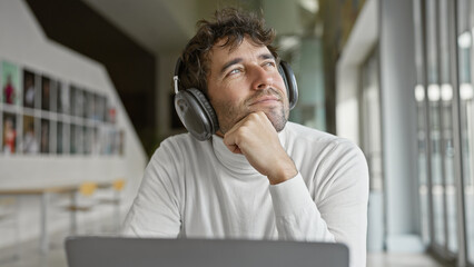 Pensive hispanic man with beard and headphones using laptop in modern office interior