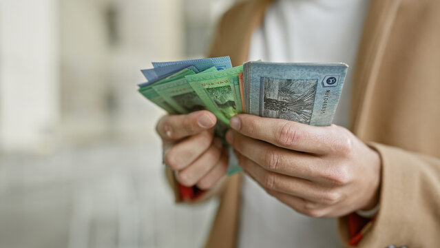 Young hispanic man counts malaysian ringgit on a busy city street, evoking themes of travel and finance.