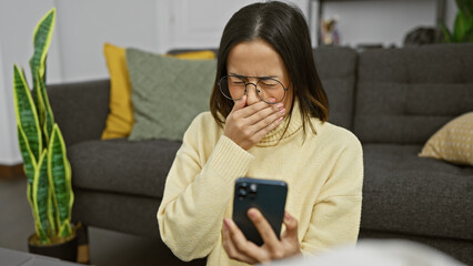 A surprised hispanic woman reads a message on her phone, sitting on a couch indoors with decor background.