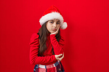 Adorable hispanic girl, bored and tired, pondering her depression issues, wearing a christmas hat. over red, isolated background with crossed arms.