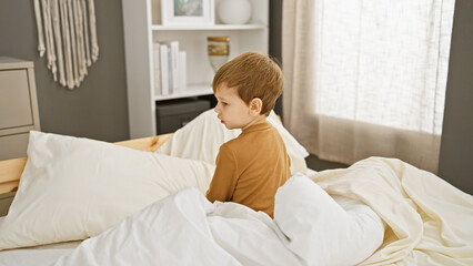 A serene caucasian toddler boy sits thoughtfully in bed, surrounded by a cozy bedroom interior.