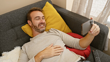 Hispanic man relaxing and taking selfie on couch with phone in home interior.