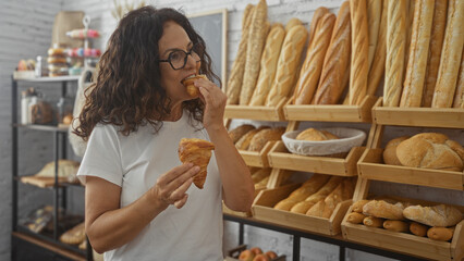 Middle-aged woman enjoying a croissant in a bakery filled with various breads and pastries, displaying a tempting and cozy indoor atmosphere.