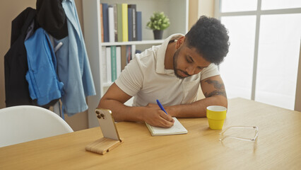 Focused man writing in notebook at home desk with smartphone, eyeglasses, and cup in view