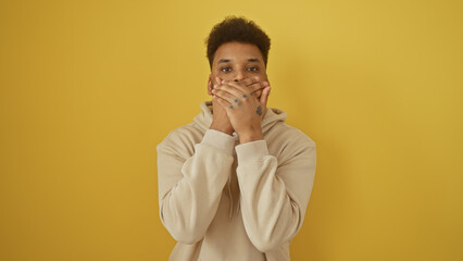 Portrait of a surprised african american man covering mouth with hands against yellow background.