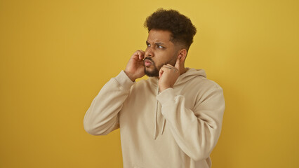 Adult black man covers ears against a solid yellow background, expressing annoyance or discomfort...
