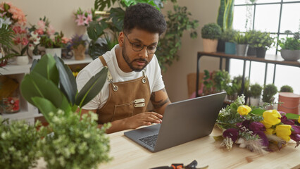 African american man working on laptop in a flower shop surrounded by fresh blossoms and greenery. - Powered by Adobe