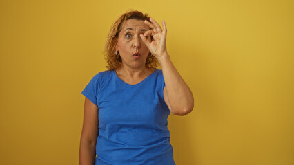 A mature hispanic woman with curly hair wearing a blue shirt makes an expressive gesture over a yellow background.