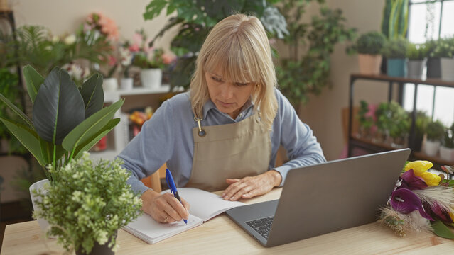 Blonde woman florist writing in notebook at a flower shop with laptop and plants in the background.