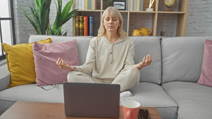Mature blonde woman meditating in a cozy living room with colorful cushions on a sofa, earphones,...