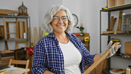 A cheerful mature woman in a woodworking workshop surrounded by tools and wooden furniture.