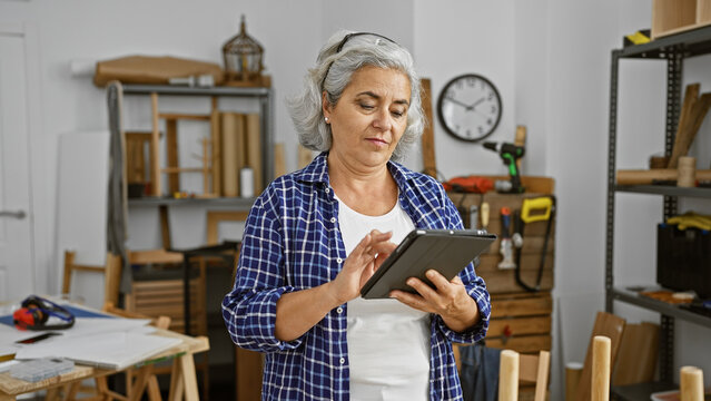 Mature woman in a workshop focused on a tablet surrounded by woodworking tools and furniture projects.