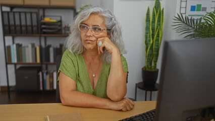 Elderly woman with grey hair and glasses sitting thoughtfully at a desk in an office room with bookshelves and plants in the background.