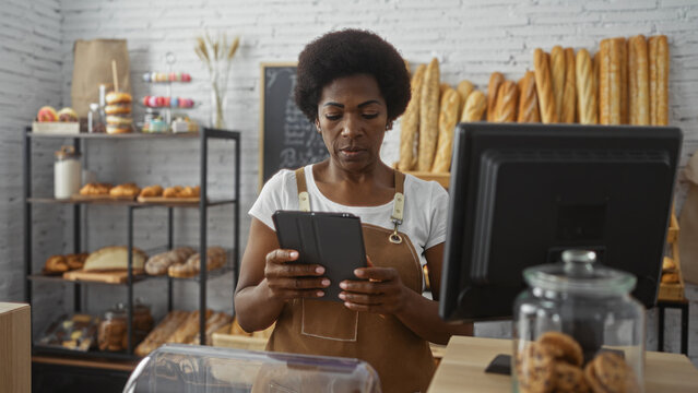 Woman working in a bakery with curly hair using a tablet in an indoor shop surrounded by bread, pastries, and a cash register - Powered by Adobe