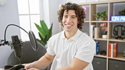 Handsome hispanic man with curly hair smiling in a radio studio, wearing a white polo shirt.
