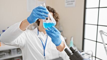 Hispanic man examines a flask with blue liquid in a bright laboratory setting, wearing protective gloves and lab coat.