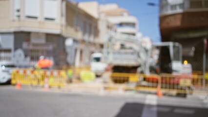 Blurred scene of construction workers and machinery outdoors with buildings in the background on a sunny day, highlighting safety barriers and street cones.