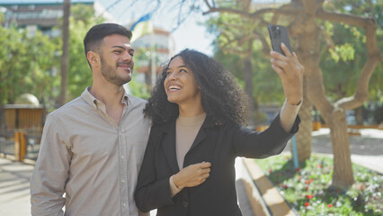 A happy couple takes a smiling selfie together in a sunny park with greenery.